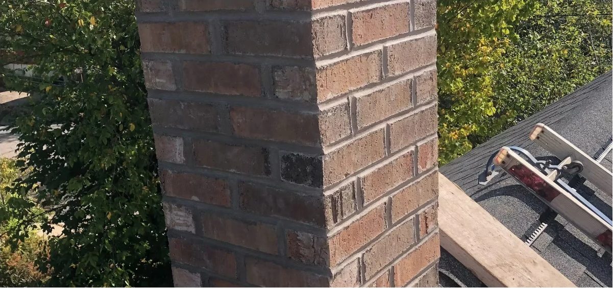 Red brick chimney with freshly repointed grey mortar joints, roof jacks and a ladder beside it